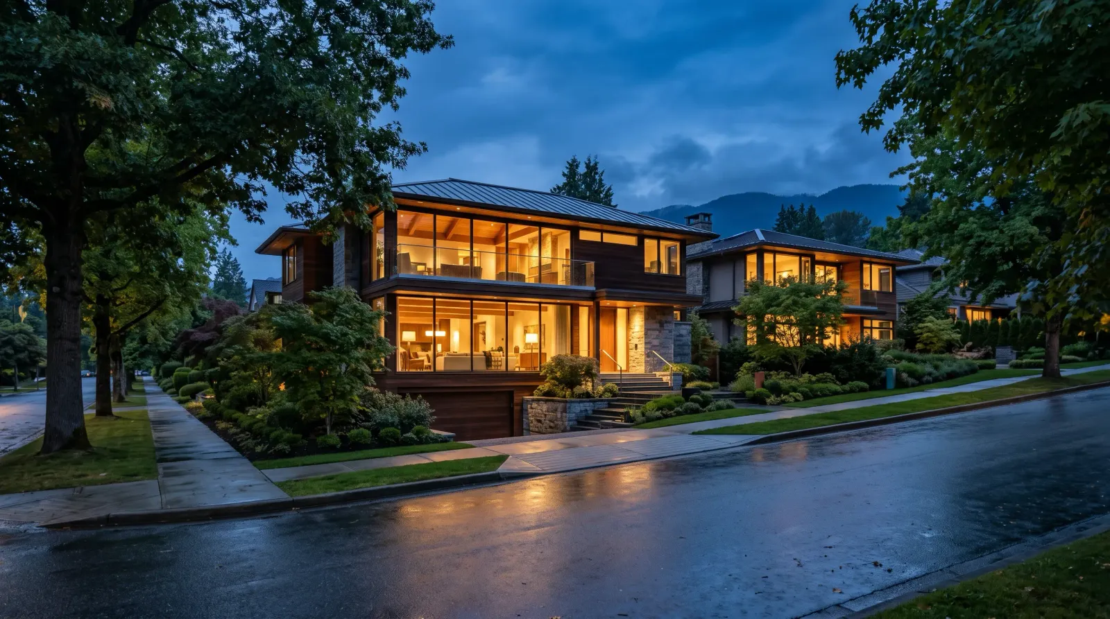 Modern West Coast homes on a tree-lined Vancouver West Side street at blue hour, warm interior lights reflecting on wet pavement with the North Shore mountains behind